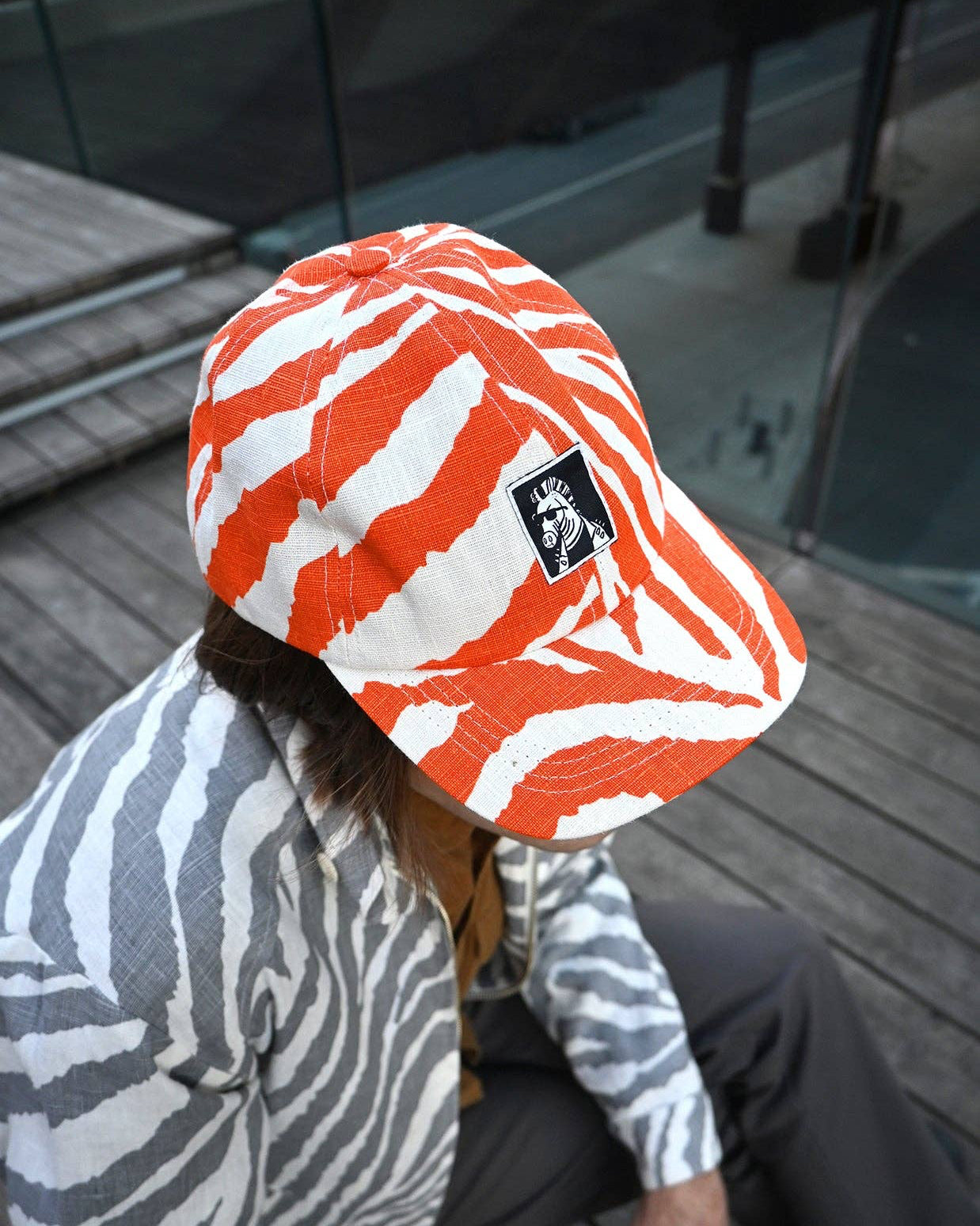 Person wearing an orange and white zebra patterned baseball hat with a zebra sitting on a wooden deck.
