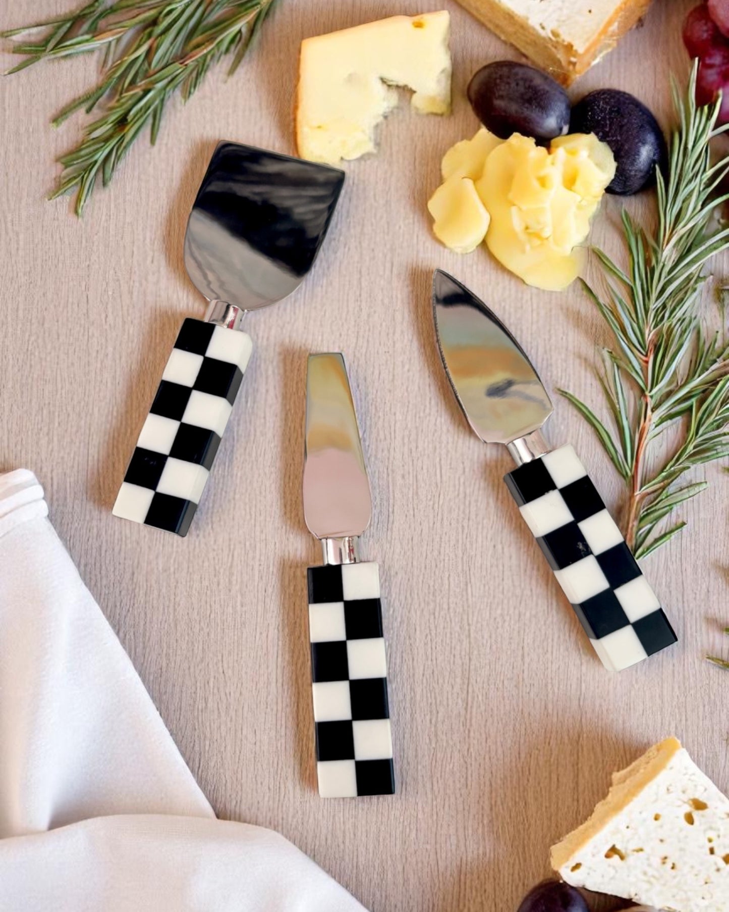 A set of cheese knives with black and white checkered handles, displayed alongside pieces of cheese and crackers.