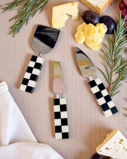 A set of cheese knives with black and white checkered handles, displayed alongside pieces of cheese and crackers.