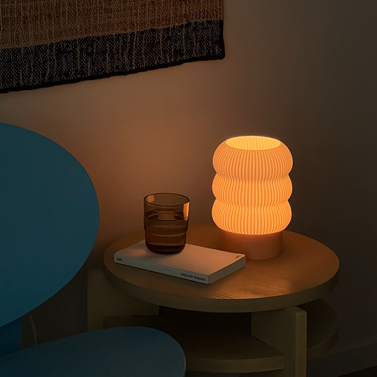 A pleated, matte finish table lamp in an orange color, placed on a wooden table with a glass of liquid and a book, with a patterned wall decor in the background.
