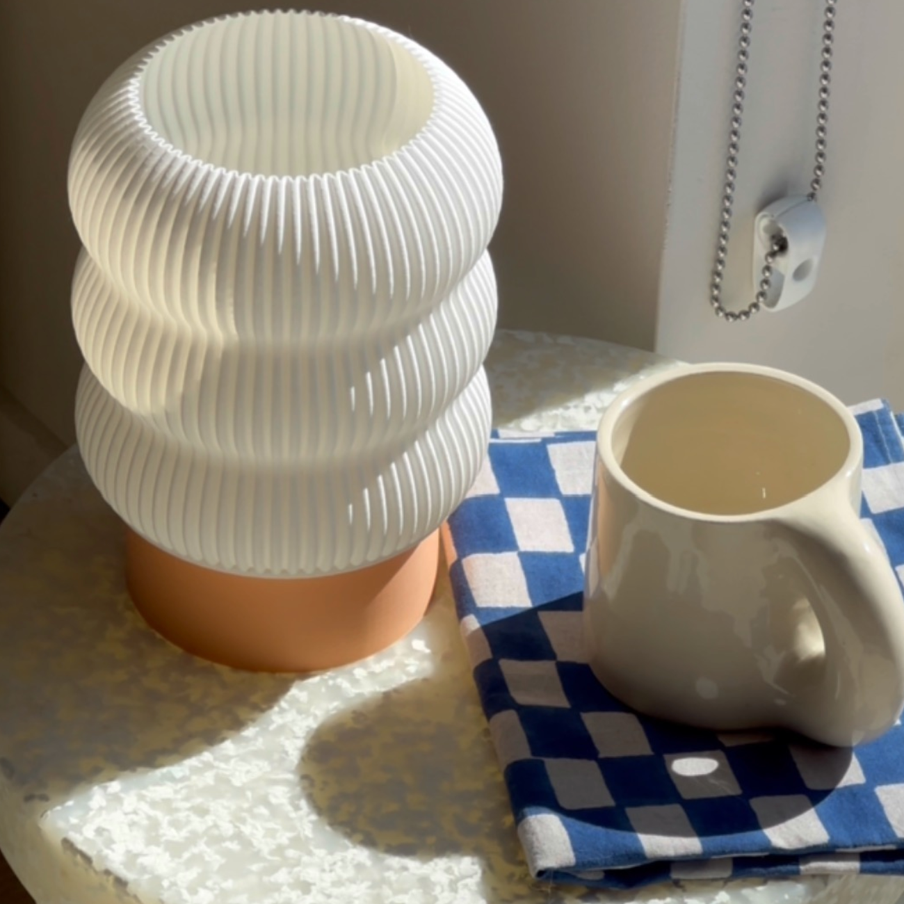 Pleated table lamp with orange base styled on a table with an ivory mug and a checkerboard napkin