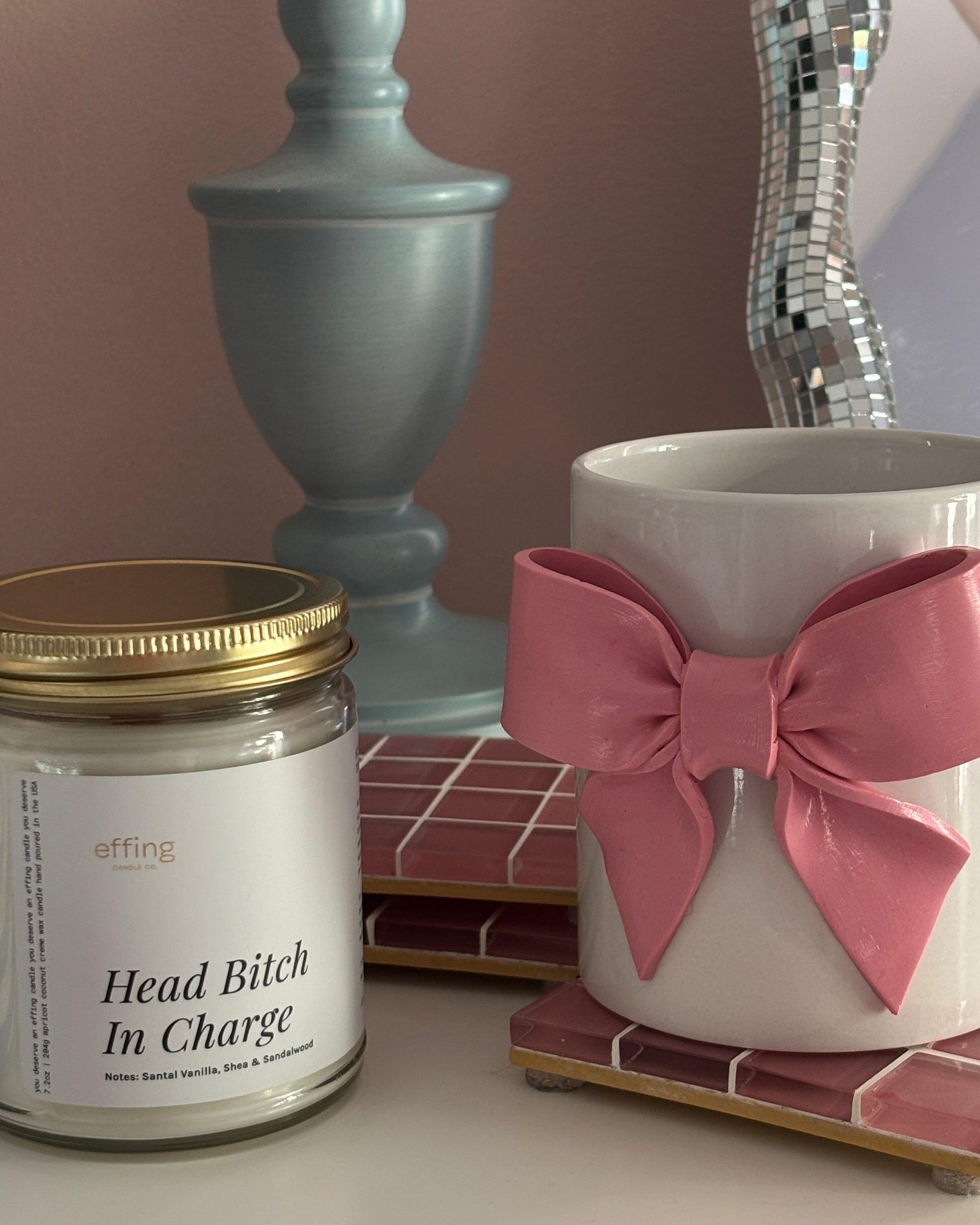 Candle labeled 'Head Bitch In Charge' next to a mug with a pink bow on a surface.