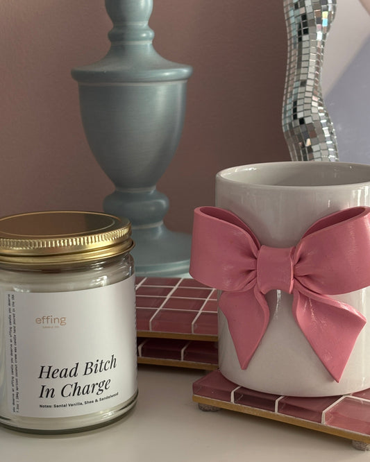 Candle labeled 'Head Bitch In Charge' next to a mug with a pink bow on a surface.